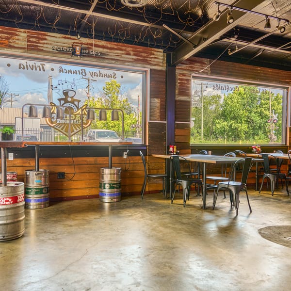 Rustic brewery interior with large windows, metal tables, concrete floor, and beer kegs.