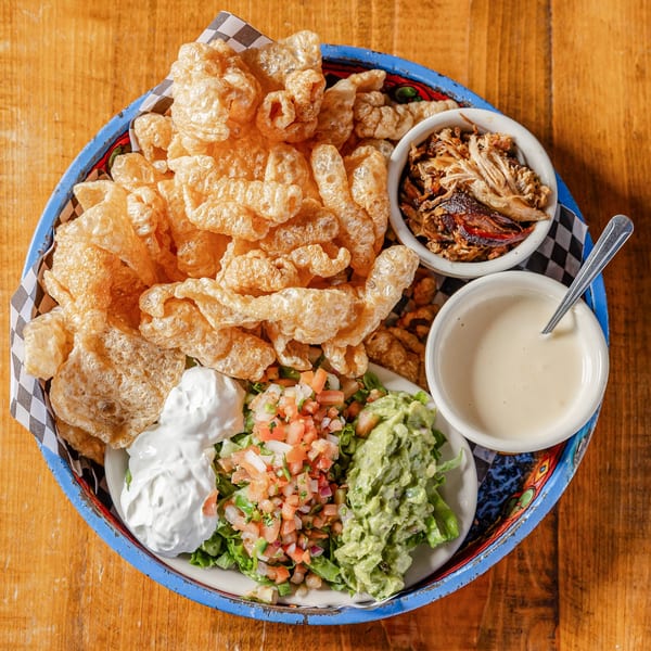 Appetizer platter with crispy chicharrones, pulled pork, guacamole, pico de gallo, and dips on a wooden table.