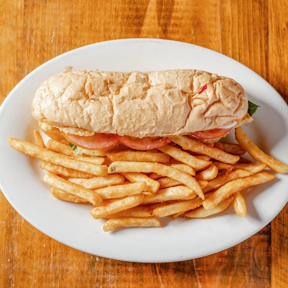 Long sandwich with tomatoes and lettuce, served with golden french fries on a white plate.
