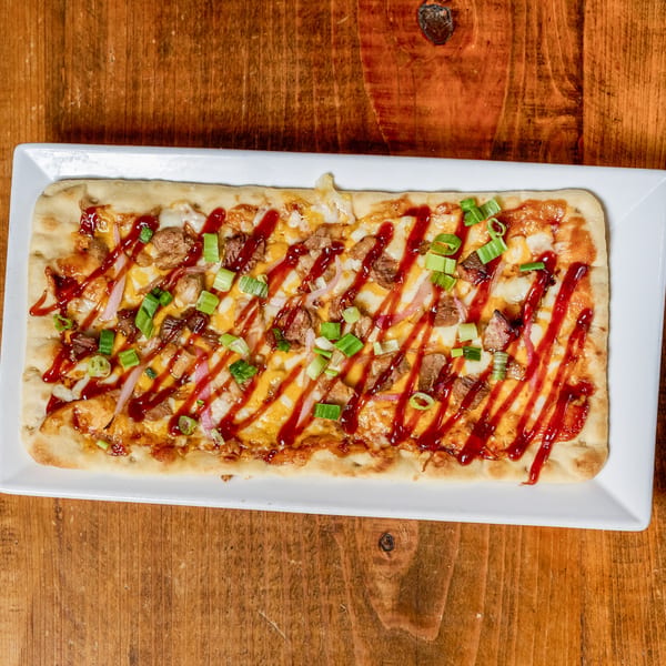 Rectangular BBQ flatbread pizza with meat, cheese, red onion, green onion, and sauce drizzles on a white plate.