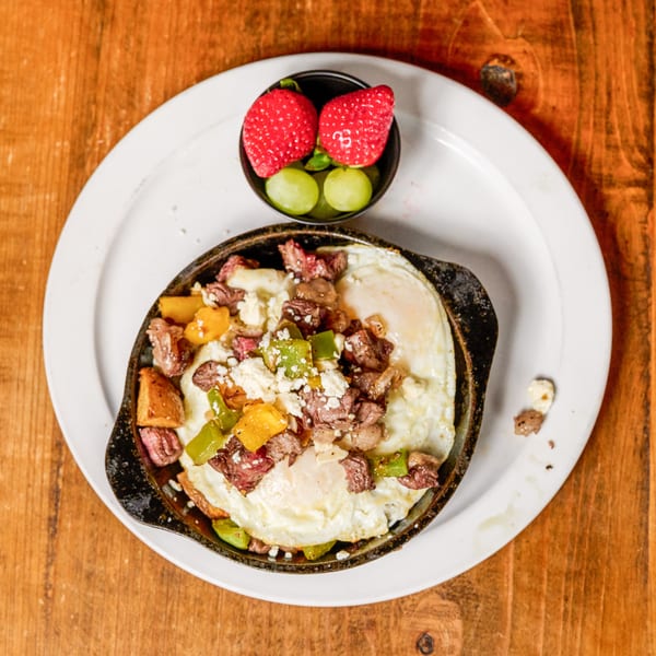 Skillet breakfast with fried eggs, steak, peppers, and cheese, served with a side of fruit.
