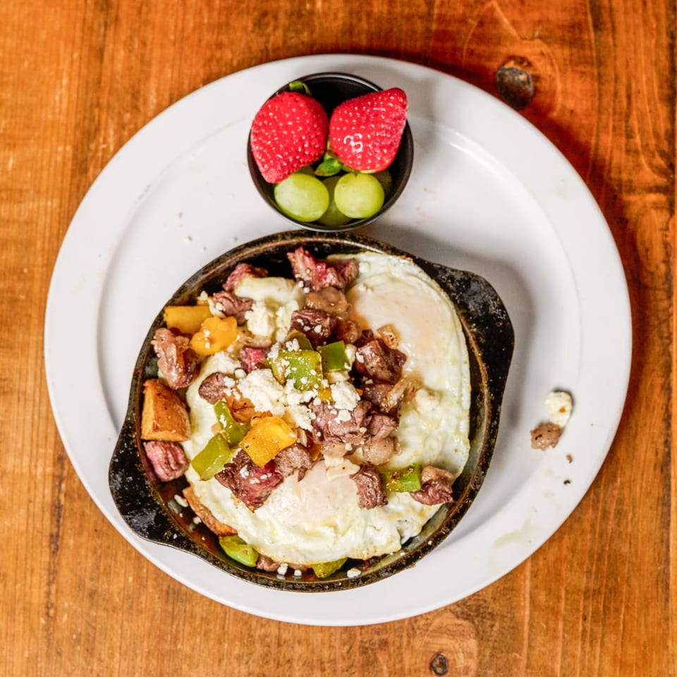 Skillet breakfast with fried eggs, steak, peppers, and cheese, served with a side of fruit.